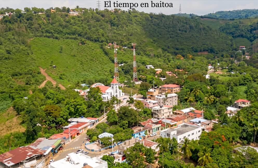 Vista aérea de alta definición del pueblo de Baitoa, República Dominicana, que muestra una iglesia blanca con techo rojo, dos torres de comunicación y un exuberante paisaje de colinas verdes. "El tiempo en baitoa"
