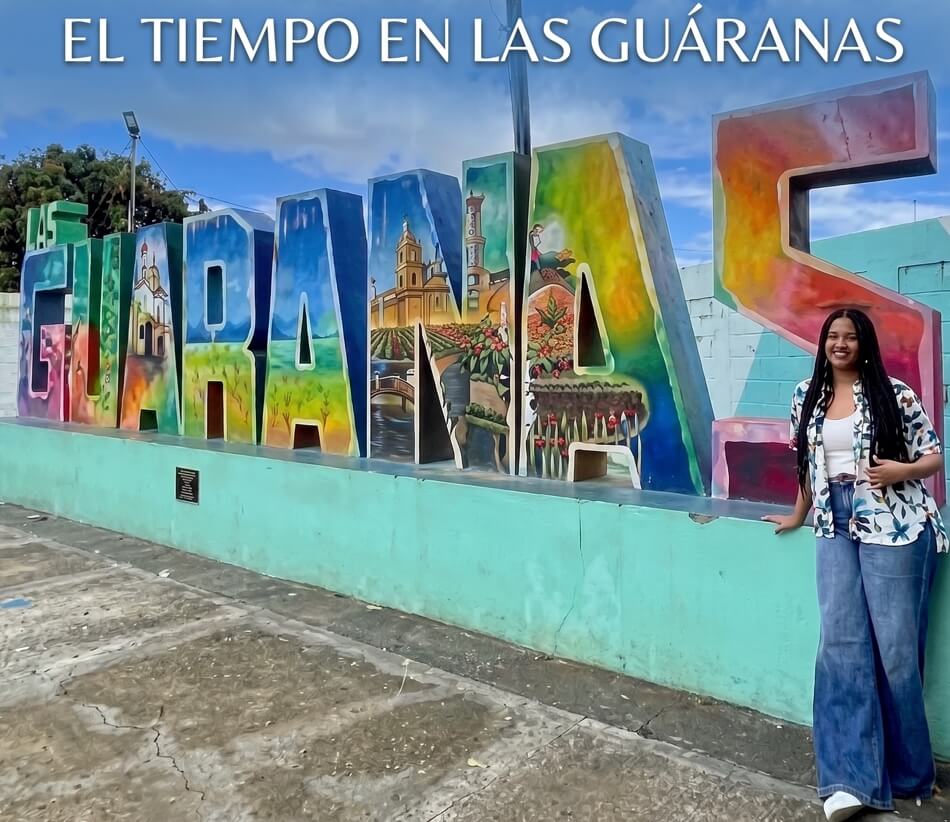 Un gran letrero corpóreo colorido que deletrea 'Las GUÁRANAS' RD con murales detallados en su interior, bajo un cielo nublado. Gloria Gonzalez "La Tati" sonriente con una camisa floral y jeans anchos posa al lado del letrero sobre un muro verde azulado."